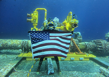 Diver underwater with United States Flag in front of some yellow subsea equipment