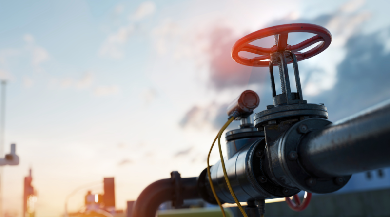 Close-up of a natural gas pipeline valve with an industrial facility in the background.