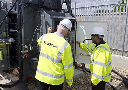 Two Power On workers in yellow high visibility jackets checking a substation