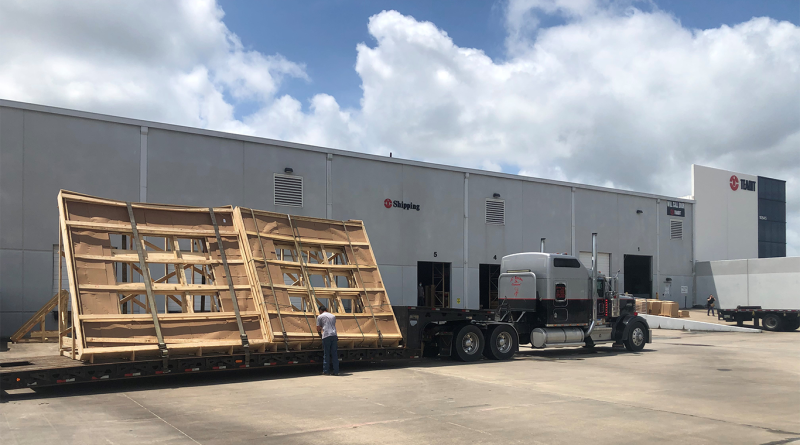Flatbed truck parked outside a warehouse loading dock carrying two large wooden shipping crates secured with straps, with workers and an industrial distribution center in the background under a partly cloudy sky.