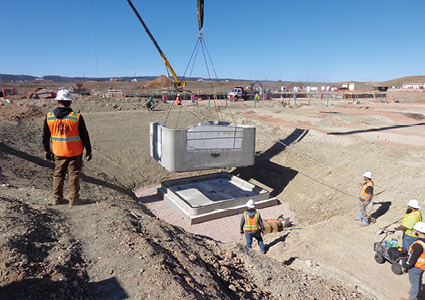 a construction crew using a crane to lower and install a precast concrete utility vault or manhole structure into an excavated pit