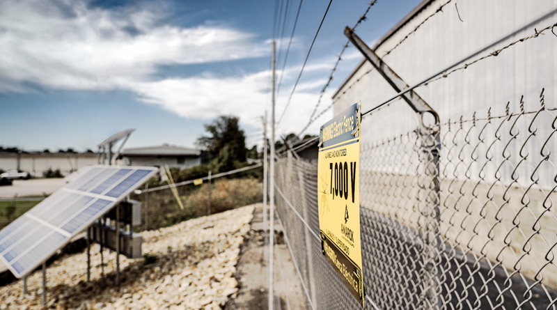 Electrified perimeter security fence with warning signage and solar panels protecting an industrial site.