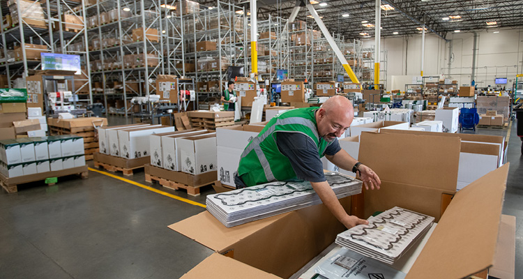a worker in a green vest packing stacks of gaskets into cardboard boxes inside a warehouse