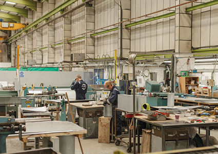 the interior of a light-industrial or metal/joinery fabrication workshop with workers at assembly tables and machinery under a high-truss steel roof