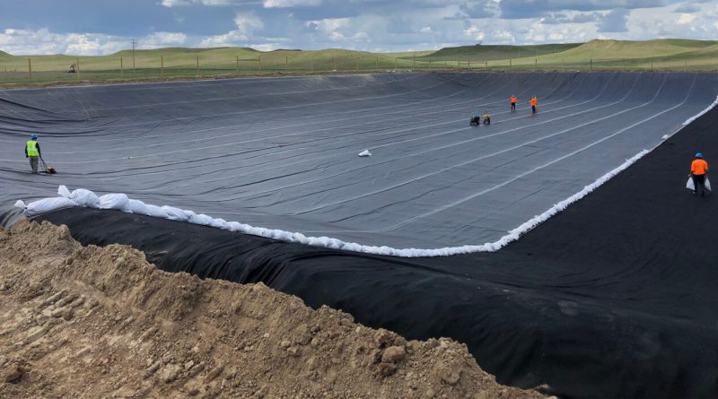 Workers installing a large black geomembrane liner across an excavated containment basin to support soil stabilisation and erosion control.